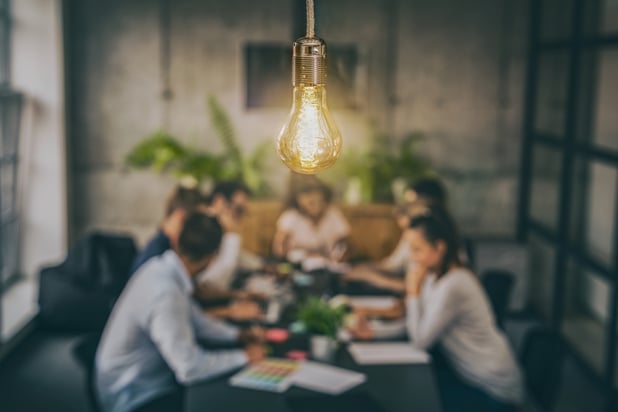 Employees experience collaboration at a conference table as a lightbulb hangs in the foreground, symbolizing inspiration